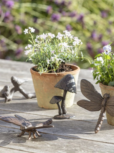 A rustic garden table styled with small terracotta pots of flowering plants, surrounded by decorative metal garden ornaments including a dragonfly, lizard and mushroom in soft natural sunlight.