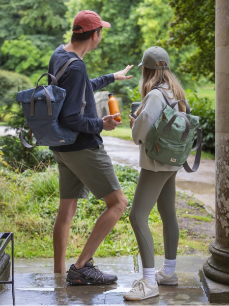 Two people wearing backpacks stand outdoors near a stone structure, looking out over a green, wooded landscape, with one gesturing ahead while holding a water bottle