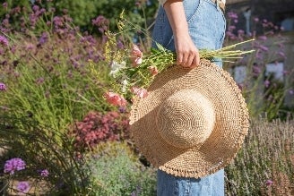Hats - Woman wearing a straw sun hat with a tan band, standing near a greenhouse surrounded by greenery.