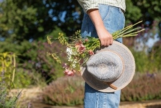 Hats - Woman wearing a straw sun hat with a tan band, standing near a greenhouse surrounded by greenery.