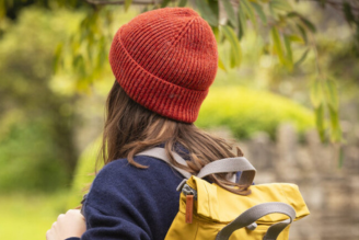 Hats - Woman wearing a straw sun hat with a tan band, standing near a greenhouse surrounded by greenery.