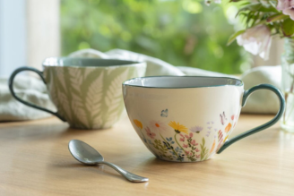A green floral ceramic tableware set displayed on a wooden shelf against a muted green wall. The set includes a large jug filled with white flowers, two mugs, a bowl, a small jug, and an espresso cup, all decorated with delicate white and red flowers and 