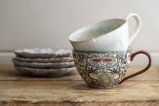 A green floral ceramic tableware set displayed on a wooden shelf against a muted green wall. The set includes a large jug filled with white flowers, two mugs, a bowl, a small jug, and an espresso cup, all decorated with delicate white and red flowers and 