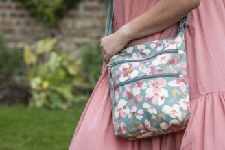 Bags - Woman wearing a National Trust crossbody bag with a colourful floral print, standing in a bright, leafy setting.