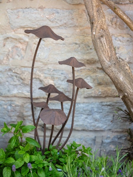 A rustic garden table styled with small terracotta pots of flowering plants, surrounded by decorative metal garden ornaments including a dragonfly, lizard and mushroom in soft natural sunlight.
