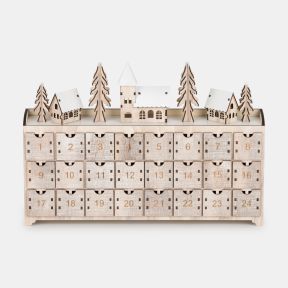 Wooden advent calendar featuring 24 numbered pull-out drawers, topped with a winter village scene including snow-covered houses, pine trees, and a central church building in a natural wood finish.
