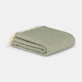 Close-up detail of a sage green woven throw blanket featuring a repeating chevron-inspired wave pattern and soft brushed finish. A row of thick cream tasselled fringe runs across the blanket, contrasting with the green fabric and highlighting the throw's