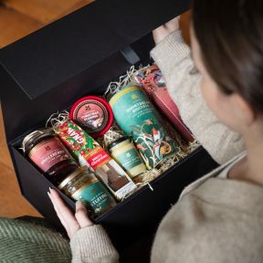 Taste of Christmas food hamper gift set presented in a black keepsake box, featuring Christmas pudding, shortbread biscuits, jingleberry jam, stuffing chutney, brandy butter and hot chocolate, arranged in decorative shredded paper packaging.