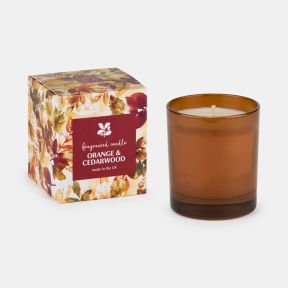 Orange & Cedarwood fragranced candle in an amber glass holder beside a matching decorative box featuring an autumn leaf design and burgundy label, displayed on a light background.