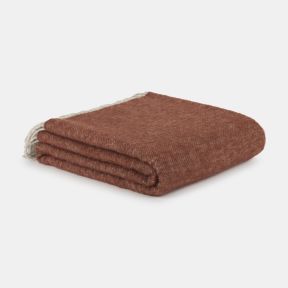 Folded rust-orange throw blanket with a soft, cosy texture, neatly stacked on a light grey background. The warm-toned fabric features a subtle woven finish, with cream fringe tassels visible along one edge for added decorative charm.