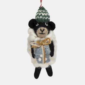 Felt sheep Christmas hanging decoration shown in a close-up view, featuring a fluffy white wool-effect coat, black face and legs, a green knitted bobble hat with white patterned detailing, and a pale blue gift wrapped with twine, suspended from a red and 