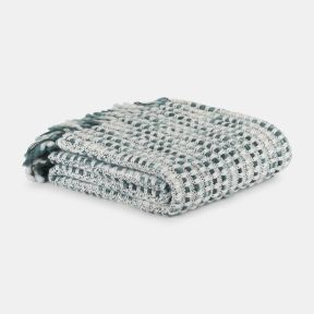 Folded teal and white woven throw blanket with a textured basket-weave pattern, neatly stacked on a light grey background. Soft fabric loops and fringe details are visible, highlighting the blanket’s cosy, handcrafted design.