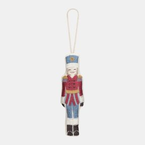 Embroidered nutcracker soldier Christmas hanging decoration featuring a red military-style jacket, blue trousers, white hat with gold star detail, and stitched facial features, finished with decorative embroidery and a cream hanging loop.