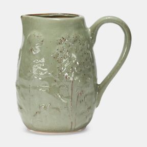 Sage green ceramic jug with a glossy glazed finish and embossed meadow-inspired floral design. The handcrafted-style pitcher features a curved handle, pouring spout and subtle speckled detailing, shown against a plain light grey background.