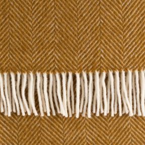 Close-up of a mustard yellow throw blanket featuring a classic herringbone weave pattern and contrasting cream fringe. The detailed view highlights the soft textured fabric, chevron design and neatly finished tasselled edge.