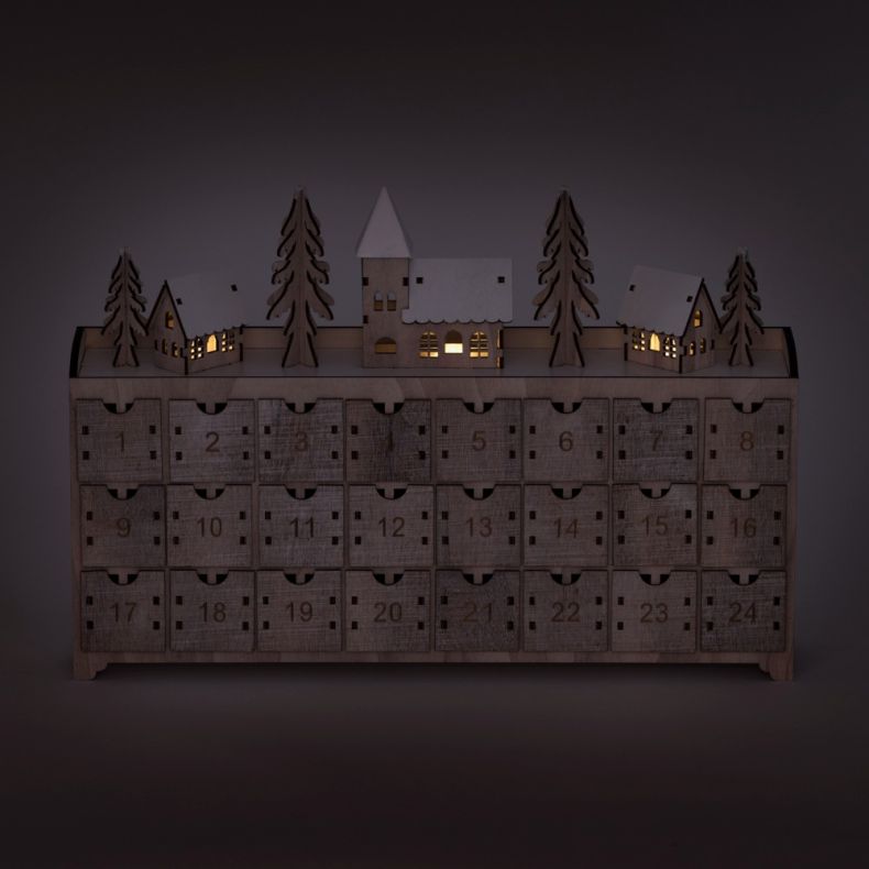 Wooden advent calendar with 24 numbered drawers and an illuminated winter village scene, featuring snow-covered houses, pine trees, and a central church building with warm glowing LED lights in a natural wood finish.