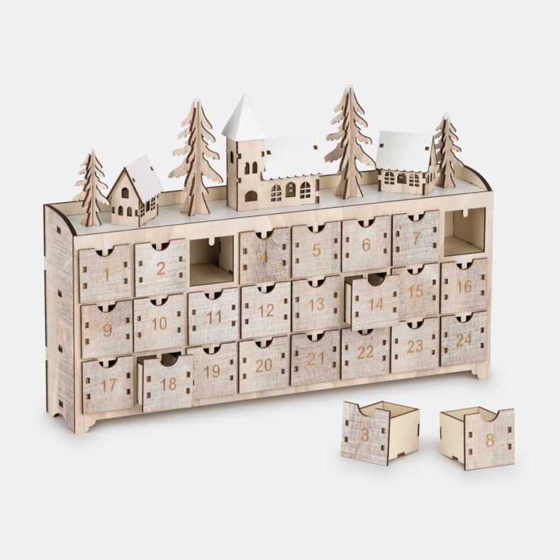 Wooden advent calendar shown from an angled view, featuring 24 numbered pull-out drawers with several drawers removed to reveal storage compartments, topped with a winter village scene including snow-covered houses, pine trees, and a central church buildi