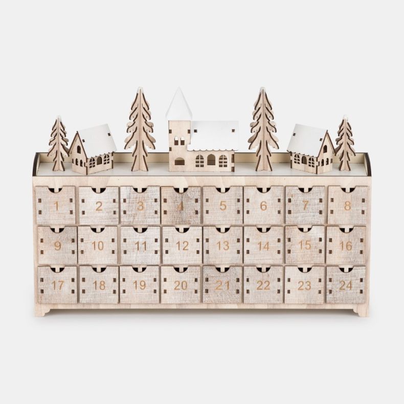 Wooden advent calendar featuring 24 numbered pull-out drawers, topped with a winter village scene including snow-covered houses, pine trees, and a central church building in a natural wood finish.