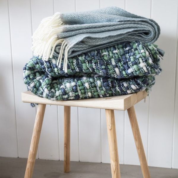 Stack of folded throws on a wooden stool, including a green, blue and ivory chunky knit recycled throw and a pale blue fringed throw.