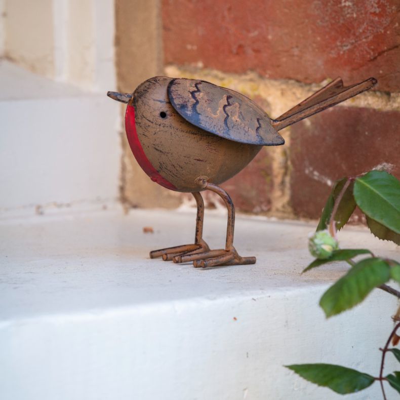 Metal robin ornament with an antique copper-effect finish, featuring a round body, red breast detail, engraved feather markings, wire legs and tail, and a freestanding design displayed on a windowsill against a brick wall background.