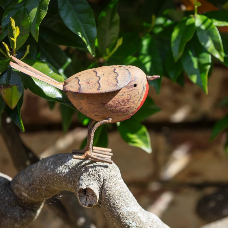 Metal robin garden ornament perched on a tree branch, featuring an antique copper-effect finish, red breast detail, engraved feather markings, wire legs and tail, and a freestanding decorative design displayed among green foliage outdoors.