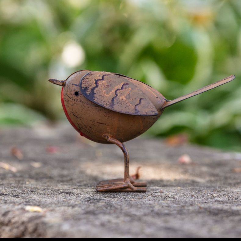 Metal robin ornament with an antique copper-effect finish, featuring a round body, red breast detail, engraved feather markings, wire legs and tail, and a freestanding design displayed on a windowsill against a brick wall background.