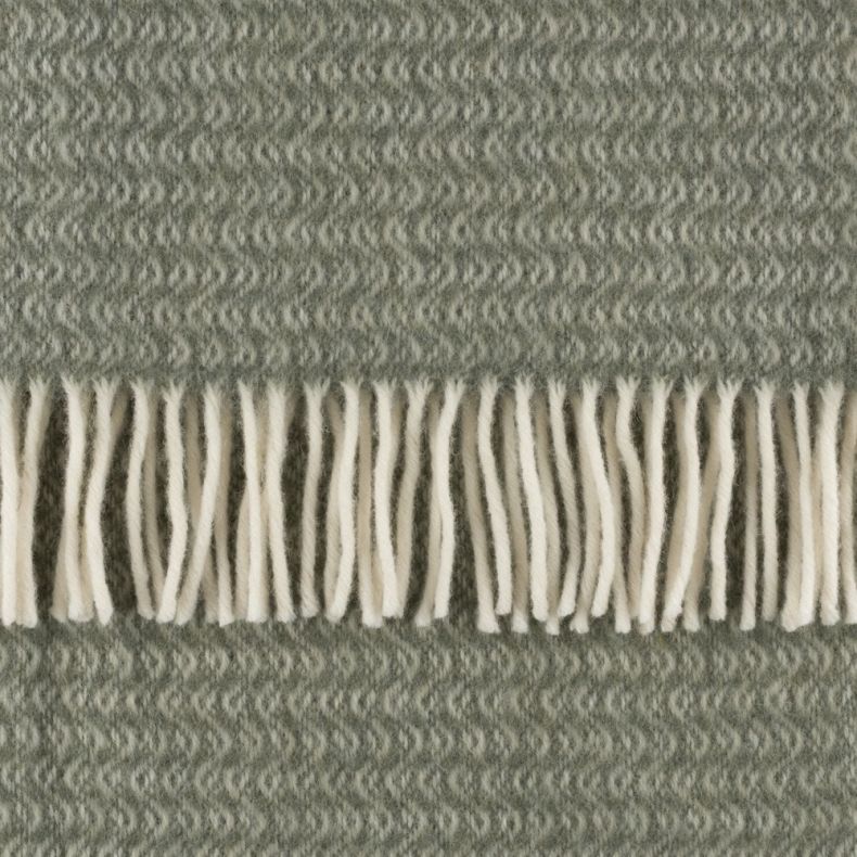 Close-up detail of a sage green woven throw blanket featuring a repeating chevron-inspired wave pattern and soft brushed finish. A row of thick cream tasselled fringe runs across the blanket, contrasting with the green fabric and highlighting the throw's 