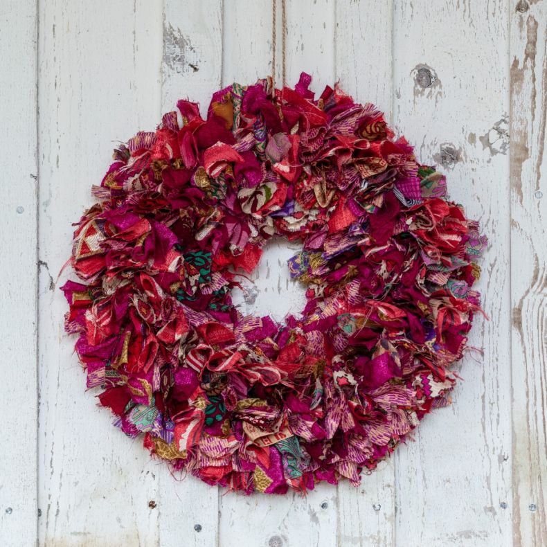 Recycled sari fabric wreath featuring vibrant pink, magenta and multicoloured textile strips, handcrafted into a circular hanging decoration and displayed against a whitewashed wooden backdrop.