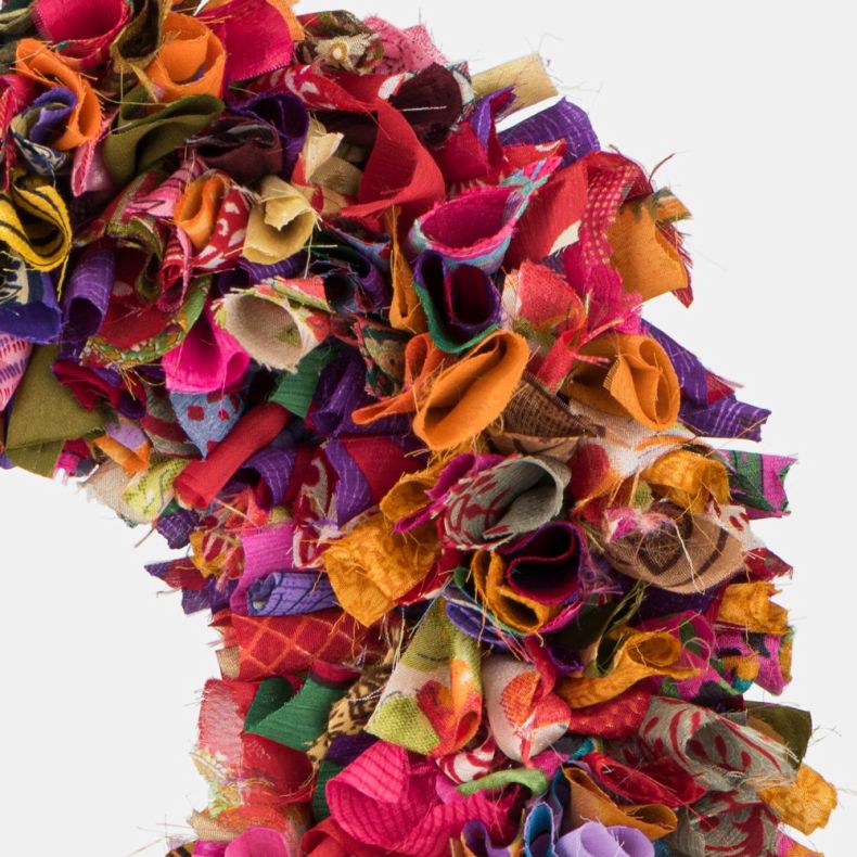 Multicoloured recycled fabric wreath close-up, highlighting densely layered strips of upcycled textiles in vibrant pink, purple, orange, red, green and blue tones, with varied printed patterns and a richly textured handcrafted finish.