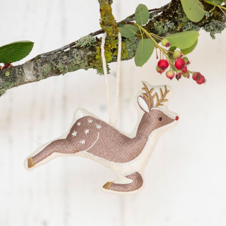 Organic cotton reindeer Christmas hanging decoration featuring an embroidered brown and cream reindeer design with gold antlers, white spotted detailing, gold hoof accents, and a natural hanging cord, displayed on a moss-covered branch with red winter ber