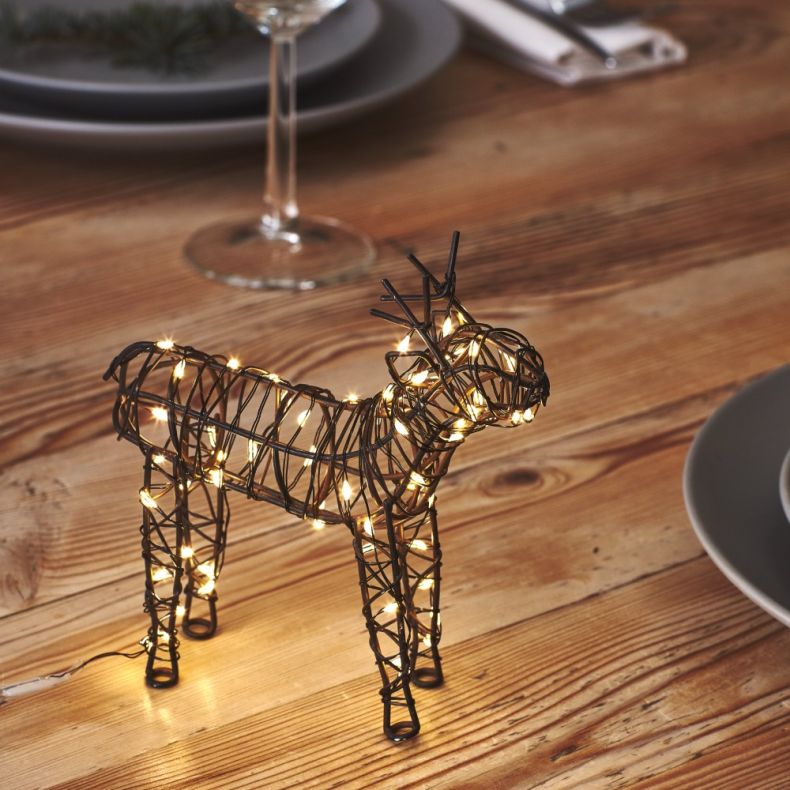 Wire reindeer Christmas decoration wrapped with warm white LED lights, featuring a black metal frame in a standing pose, displayed on a wooden tabletop as a festive indoor seasonal ornament.