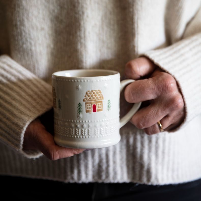 Cream ceramic mug with an embossed festive pattern, featuring a gingerbread house motif with pine tree accents in soft brown, green and red tones, shown held in both hands with a cosy cream knitted jumper.