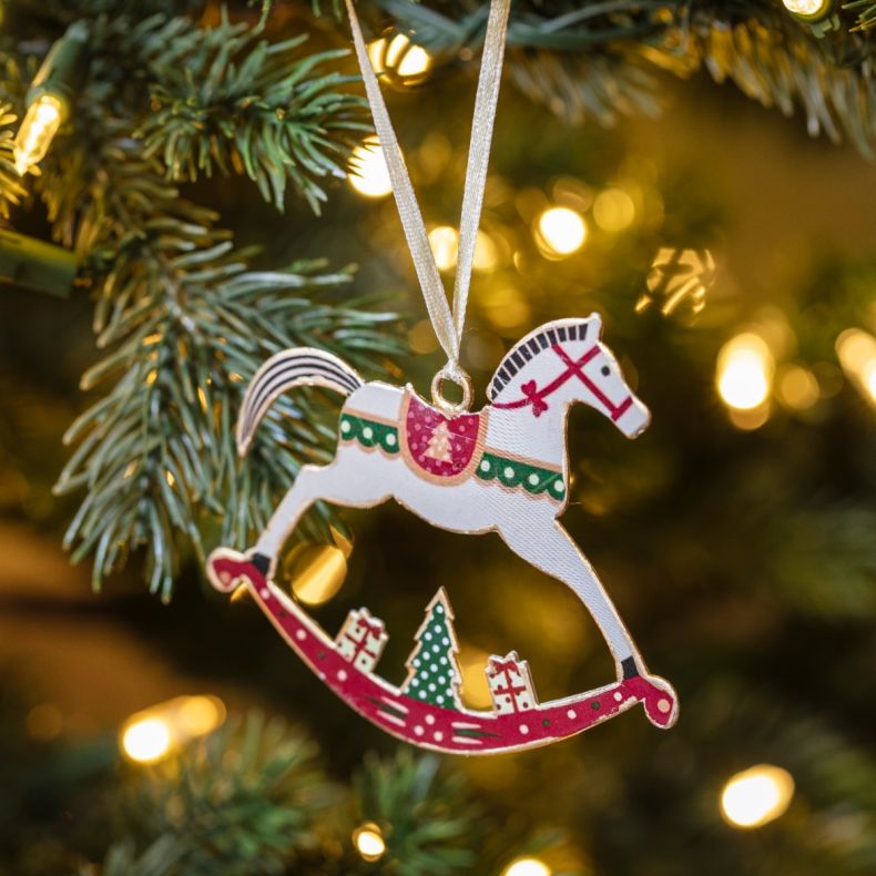 Wooden rocking horse Christmas hanging decoration displayed on a Christmas tree, featuring a cream horse with red bridle and saddle details, green scalloped trim, a festive Christmas tree motif, gift illustrations on the curved rocking base, gold-tone edg