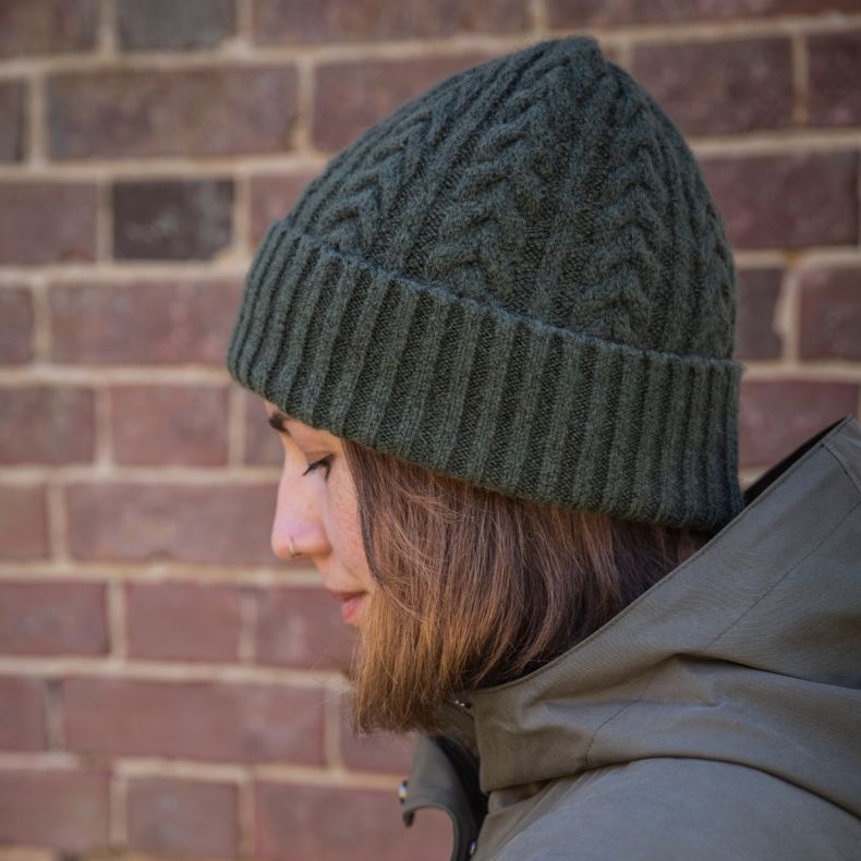 Dark green cable knit beanie hat with ribbed turn-up cuff, shown worn outdoors in side profile view against a brick wall background