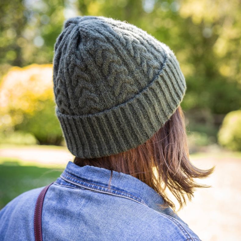 Dark green cable knit beanie hat with ribbed turn-up cuff, shown worn outdoors from the rear, highlighting the textured knit pattern and fitted crown shape