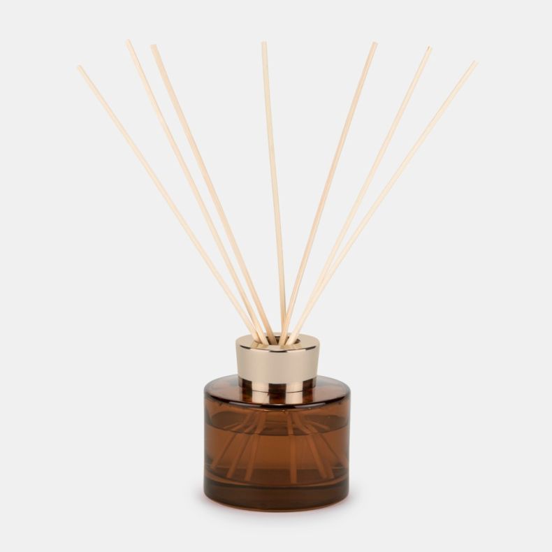 Amber glass reed diffuser with a gold-tone collar and natural reed sticks fanned out from the bottle, displayed against a light background.