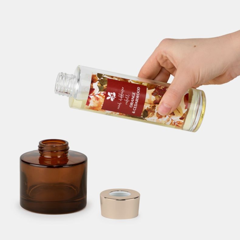 Hand pouring Orange & Cedarwood reed diffuser refill from a clear labeled bottle into an open amber glass diffuser vessel, with the gold-tone collar placed beside the bottle on a light background.