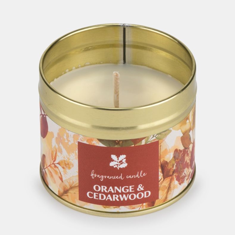 Orange & Cedarwood fragranced candle in an open gold-coloured metal tin, revealing smooth white wax and a single centred wick. The candle features autumn-inspired leaf artwork in warm orange, amber, and cream tones with a matching fragrance label on the f