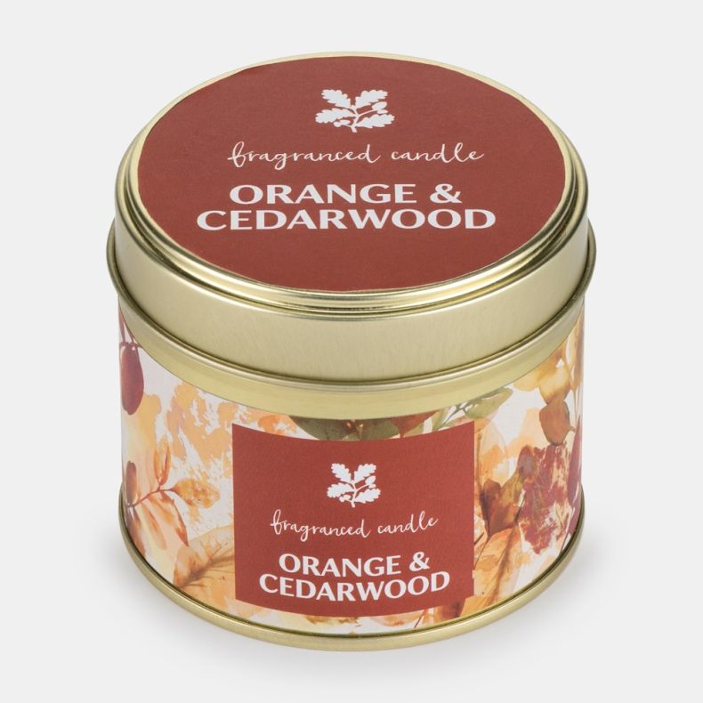 Orange & Cedarwood fragranced candle in a gold-coloured metal tin with a fitted lid. The candle features autumn-inspired leaf artwork in warm orange, amber, and cream tones, with matching front and lid labels displaying the fragrance name.