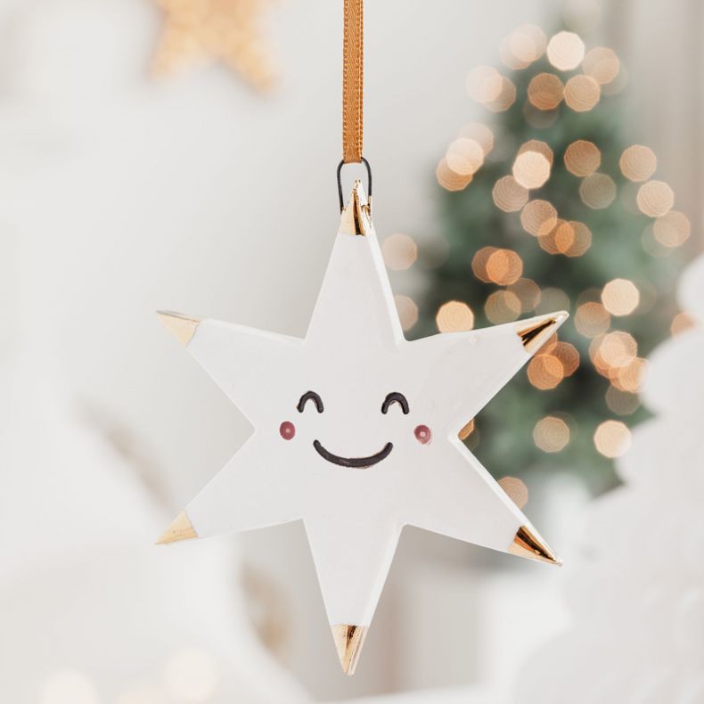 White ceramic star Christmas hanging decoration displayed in a festive setting, featuring a smiling face with pink cheek details, gold-tone tipped points, a glossy glazed finish, and a gold hanging ribbon, shown with softly blurred Christmas tree lights i