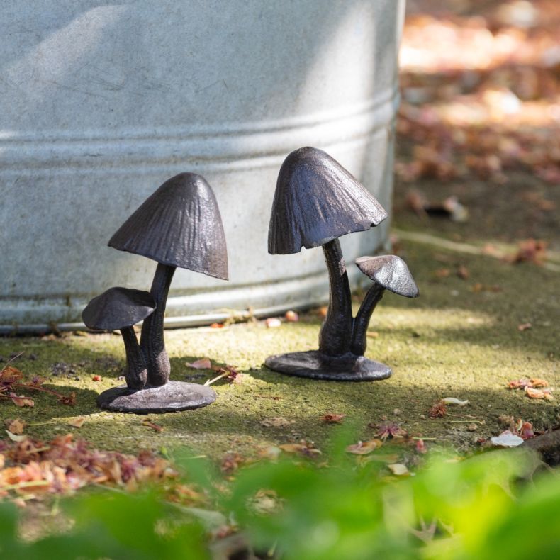 Two cast iron mushroom sculptures featuring textured stems and detailed caps, displayed on a mossy stone surface beside a galvanised metal planter, with scattered leaves and soft-focus greenery in the foreground.