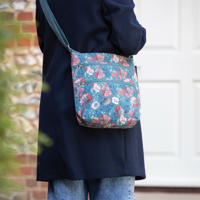 Blue floral crossbody bag worn over a navy coat, featuring an adjustable shoulder strap and two front zip pockets.