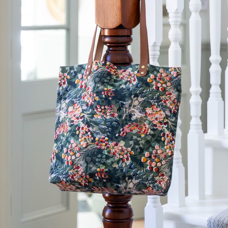 Floral print tote bag with tan shoulder straps hanging indoors, featuring an all-over botanical berry pattern in shades of green, pink and red.