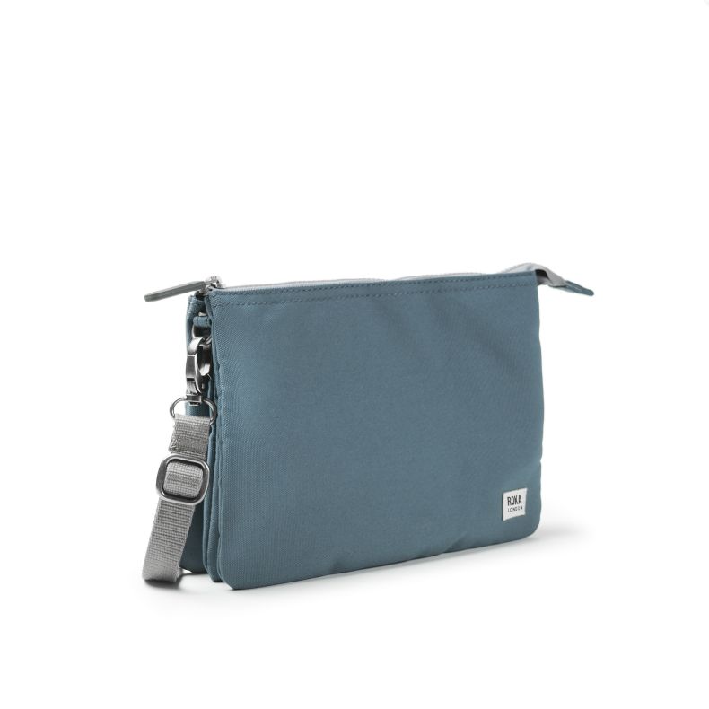 Blue-grey recycled canvas crossbody pouch shown from an angled side view, featuring a compact double-compartment design, adjustable webbing strap, zip-top closures, silver-tone hardware, and woven logo label.