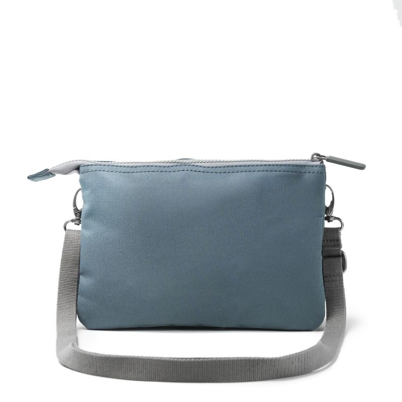Blue-grey recycled canvas crossbody pouch shown from the reverse side, featuring a clean minimalist design, adjustable webbing strap, zip-top closure, silver-tone hardware, and durable textured fabric.