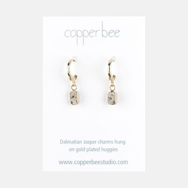 Gold-tone huggie hoop earrings with rectangular dalmatian jasper stone drop charms, displayed on a branded jewellery card featuring cream-toned natural stone with grey and black speckled detailing.