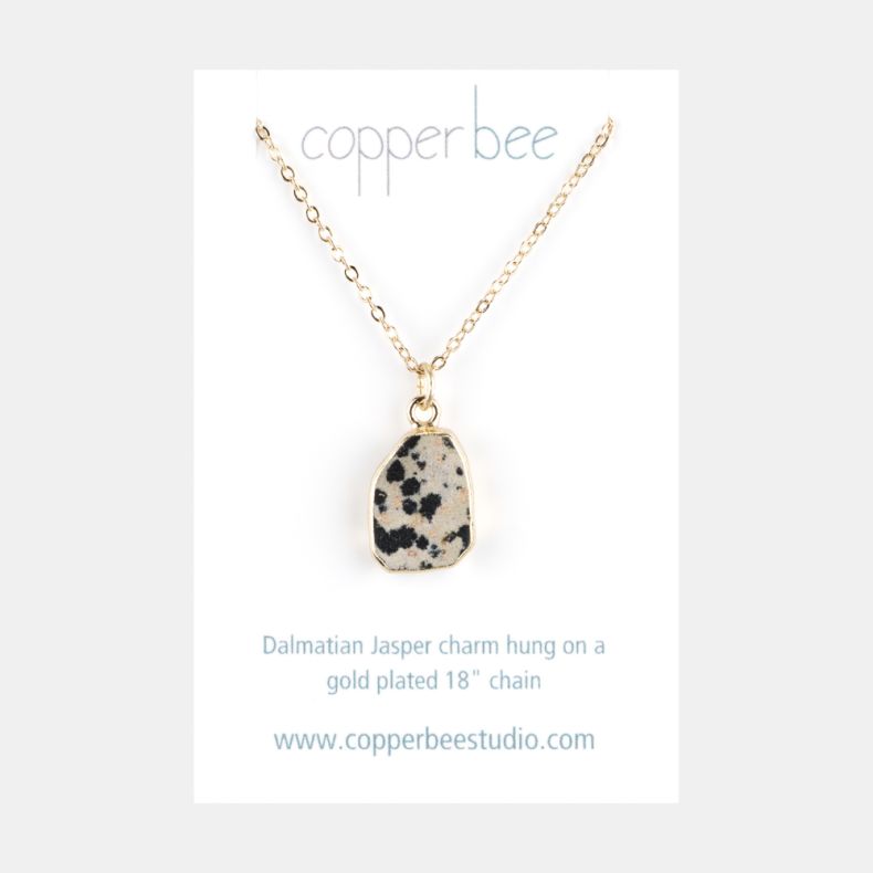 Gold-tone necklace with an irregular dalmatian jasper stone pendant on a fine link chain, displayed on a branded jewellery card featuring a cream-toned natural stone design with black speckled detailing.
