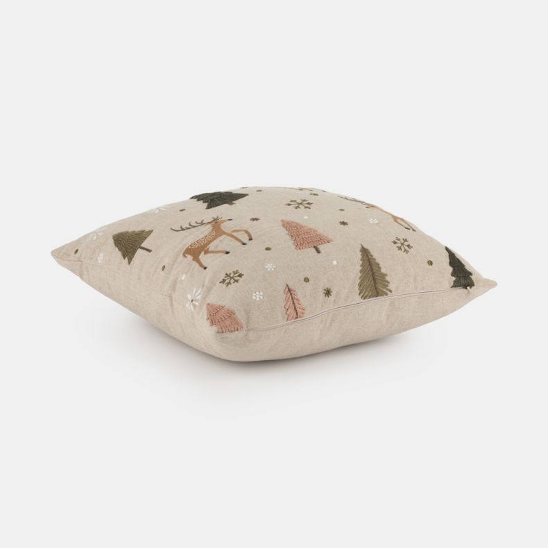 Beige embroidered cushion shown from an angled side view, featuring stag, fir tree and snowflake motifs in soft brown, green, pink and cream tones, with textured stitched detailing and a natural linen-look fabric cover