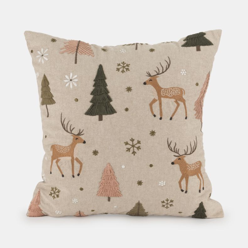 Beige embroidered cushion featuring stag, fir tree and snowflake motifs in soft brown, green, pink and cream tones, with textured stitched detailing on a natural linen-look fabric cover.