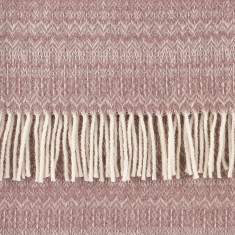 Close-up of a blush pink woven throw blanket featuring a subtle geometric and zigzag pattern with soft cream fringe tassels. The cosy fabric has a textured finish, highlighting the intricate weave and warm, inviting design.